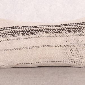 Puede incluir: Cojín rectangular beige con rayas y detalles cosidos en marrón oscuro. El cojín tiene un aspecto texturizado y tejido, con detalles de costura horizontales y verticales. El cojín está colocado sobre una superficie neutra.