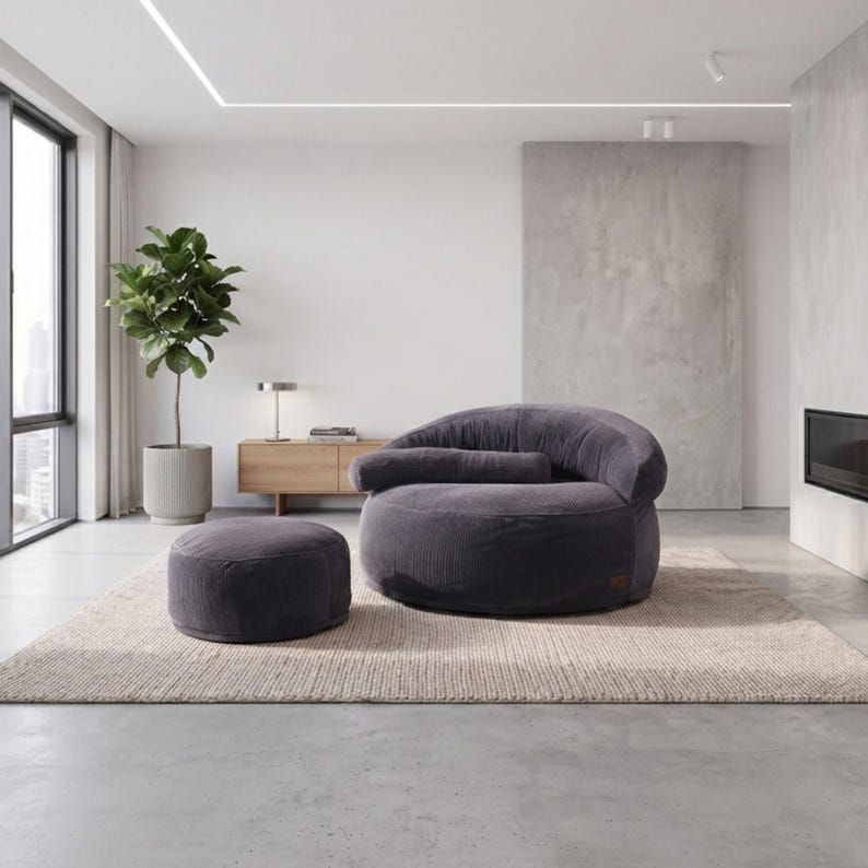 May include: A dark gray corduroy lounge chair with a rounded back and a matching cylindrical ottoman. The chair features a horizontal bolster pillow across the seat, and both pieces are upholstered in a textured, ribbed fabric with a soft finish.