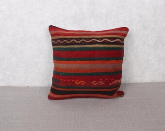May include: A rectangular pillow with a woven design. The pillow features horizontal stripes in shades of red, orange, green, and brown. The pillow has a textured appearance and is likely made of a natural fiber.
