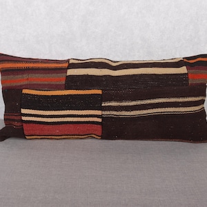 May include: Rectangular decorative pillow with a patchwork design. The pillow features various sections with horizontal stripes in shades of brown, beige, orange, and red. The pillow is placed on a gray surface.