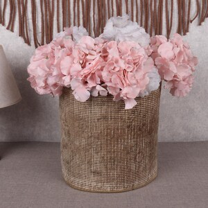 May include: A cylindrical woven basket filled with pink and white faux hydrangeas. The basket has a textured, natural appearance. The flowers are clustered together, creating a full display. A wooden lamp with a beige lampshade is on the left.