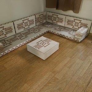 May include: A beige sectional sofa with a geometric pattern in brown, along with a matching square ottoman. The sofa is positioned in a corner, and the room has hardwood flooring. A small side table with a plant is also visible.