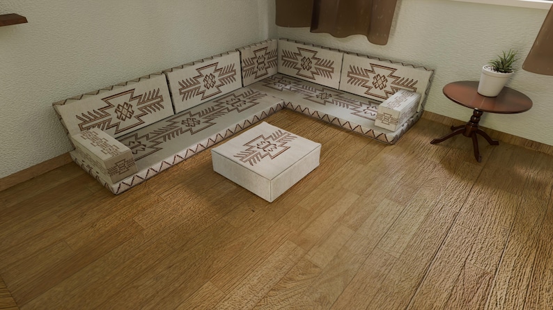 May include: A beige sectional sofa with a geometric pattern in brown, along with a matching square ottoman. The sofa is positioned in a corner, and the room has hardwood flooring. A small side table with a plant is also visible.