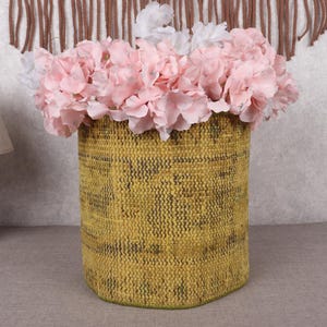 May include: A cylindrical, woven basket in shades of yellow and green, filled with pink and white artificial flowers. The basket is placed on a gray surface, with a wooden lamp and decorative wall hanging in the background.
