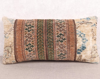 May include: Rectangular decorative pillow with a vintage, distressed look. The pillow features a central panel with green and brown geometric patterns, flanked by brown stripes and beige sections with blue accents. The pillow has a soft, textured appearance.