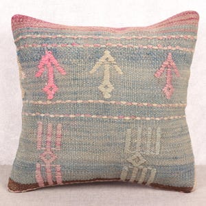 May include: A square decorative pillow with a woven texture. The pillow features a light blue background with pink and off-white arrow and geometric designs. The top and bottom edges are brown, and the top edge is pink.