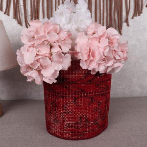 Puede incluir: Una cesta roja tejida llena de flores artificiales rosas y blancas. La cesta tiene forma cilíndrica y una superficie texturizada. Las flores están dispuestas para crear una exhibición completa y exuberante, agregando un toque de color y textura.