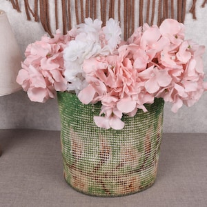 May include: A green and pink woven basket filled with pink and white flowers. The basket has a textured appearance and is cylindrical. A wooden lamp with a beige lampshade is to the left.