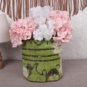 May include: A green and brown patterned vase holds pink and white flowers. The vase is cylindrical and has a woven texture. A wooden lamp with a beige lampshade is to the left.