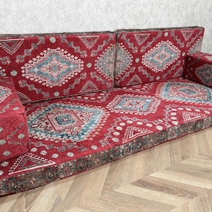 May include: A red upholstered sofa with a patterned design in shades of blue, gray, and white. The sofa has a low profile and includes a seat, back cushions, and two armrests. The fabric has a traditional, oriental rug-like pattern.