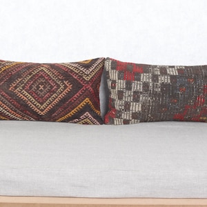 Puede incluir: Dos cojines rectangulares con intrincados patrones tejidos. Uno presenta un diseño de rombos en tonos marrones, rojos y amarillos. El otro tiene un patrón de cuadros con acentos rojos y blancos sobre un fondo oscuro.