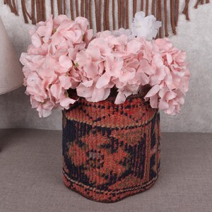 May include: A floral arrangement featuring light pink hydrangeas in a decorative, cylindrical vase. The vase has a patterned design in shades of red, black, and brown. A wooden lamp with a beige lampshade is visible on the left.