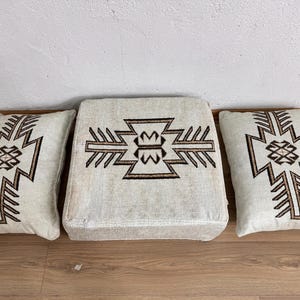 May include: A set of three beige cushions with a geometric brown design. The set includes two rectangular pillows and a square ottoman, all featuring a symmetrical pattern. The cushions are placed on a wooden floor.