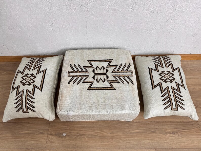 May include: A set of three beige cushions with a geometric brown design. The set includes two rectangular pillows and a square ottoman, all featuring a symmetrical pattern. The cushions are placed on a wooden floor.