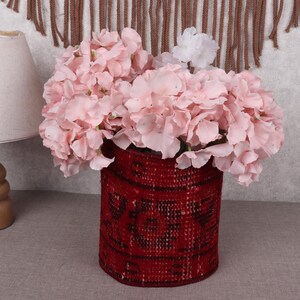 May include: A red woven vase filled with pink flowers and a single white flower. The vase has a cylindrical shape and a textured surface. A wooden lamp with a beige lampshade is to the left.