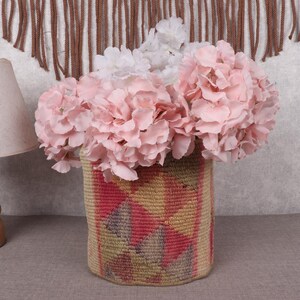 May include: A decorative woven basket with a geometric pattern, filled with pink and white flowers. A wooden lamp with a beige shade is to the left. The background includes a textured wall and a brown woven wall hanging.