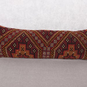 May include: Rectangular decorative pillow with a geometric pattern in shades of burgundy, orange, and gold. The pillow has a textured appearance and is placed on a neutral gray surface. The pillow's design features a repeating pattern.