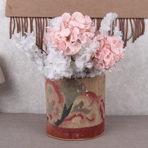 May include: A cylindrical vase with a tapestry design, filled with pink and white flowers. The vase has a red border and a beige background with a floral pattern. A wooden lamp with a beige lampshade is on the left.