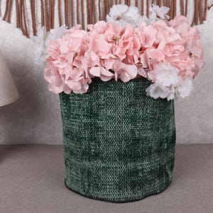 May include: A green textured vase filled with pink and white flowers. The vase has a cylindrical shape and sits on a gray surface. A wooden lamp with a beige lampshade is to the left.