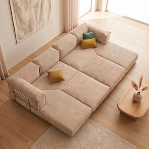 Peut inclure: Un grand canapé modulaire en velours côtelé beige au design bas, composé de plusieurs coussins d'assise et de dossier rectangulaires. Le canapé comprend un petit coussin carré bleu sarcelle et deux coussins carrés jaune moutarde.
