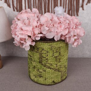 May include: A cylindrical green woven vase filled with pink hydrangea flowers. The vase has a textured pattern and is set against a neutral background. A lamp with a beige shade is visible on the left.
