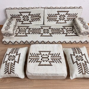 May include: A beige floor seating set with geometric brown patterns. The set includes a sofa, two armrests, two pillows, and a square ottoman. The sofa and ottoman have a solid base, while the pillows are soft and rectangular. The design is symmetrical.