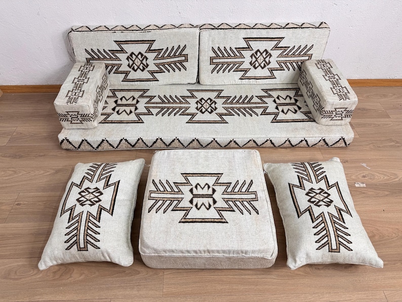 May include: A beige floor seating set with geometric brown patterns. The set includes a sofa, two armrests, two pillows, and a square ottoman. The sofa and ottoman have a solid base, while the pillows are soft and rectangular. The design is symmetrical.