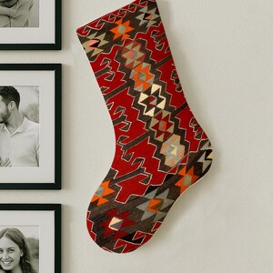 May include: A Christmas stocking with a geometric pattern in shades of red, brown, orange, and gray hangs on a white wall. The stocking has a white cuff and a loop for hanging.