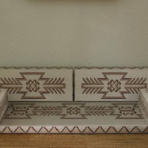 May include: A beige floor sofa with geometric brown patterns. The sofa has two back cushions and two armrests. The design is repeated on the seat and armrests. The sofa is placed on a wooden floor against a textured wall.