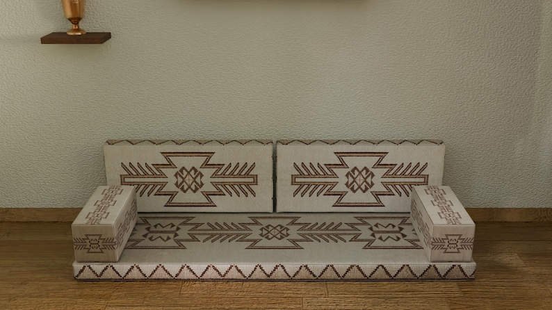 May include: A beige floor sofa with geometric brown patterns. The sofa has two back cushions and two armrests. The design is repeated on the seat and armrests. The sofa is placed on a wooden floor against a textured wall.
