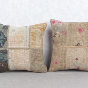 May include: Two square patchwork throw cushions in muted tones of beige, green, and pink. The cushions feature a textured fabric and a patchwork design. They are displayed on a grey surface.