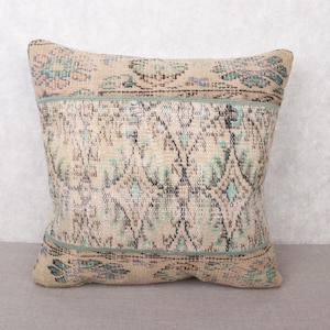 May include: Square decorative pillow with a beige, blue, and black patterned design. The pillow has a vintage, distressed look, with a faded appearance. The pillow is made of a woven fabric and has a soft texture. The pillow is placed on a gray surface.