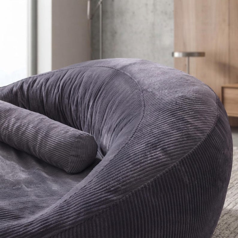 May include: A dark gray corduroy armchair featuring a curved, rounded silhouette and a matching cylindrical bolster pillow. The fabric has a distinct vertical ribbed texture that covers the entire exterior and interior of the seat.