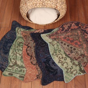 May include: A collection of patterned pet bed mats in various colors, including navy, green, and rust, arranged on a wooden floor. A woven wicker pet bed with a white cushion is positioned above the mats.