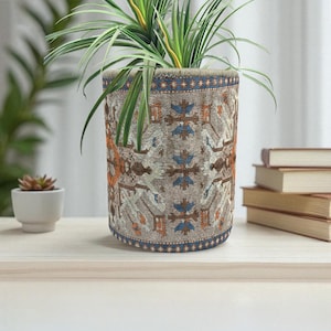 Puede incluir: Una maceta cilíndrica con un diseño de tela estampada en tonos beige, naranja y azul. La maceta contiene una planta verde con hojas largas y delgadas. La maceta está sobre una superficie de madera clara.