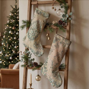 May include: Two Christmas stockings made of a light beige fabric featuring a detailed teal and blue floral pattern. Each stocking has a brown leather hanging loop at the top and is displayed hanging from the rungs of a wooden ladder.