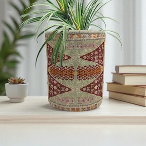 Puede incluir: Una maceta cilíndrica con un diseño de tela detallado en tonos rojos, beige y naranja. La maceta contiene una planta verde con hojas largas y delgadas. La maceta está sobre una superficie de madera clara.