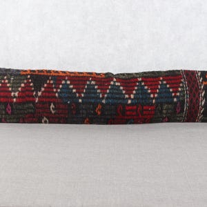 May include: A long, rectangular pillow with a woven design. The pillow features a pattern of red, blue, and black geometric shapes. The ends of the pillow are black, with an orange design on one end. The pillow is likely used for home decor.