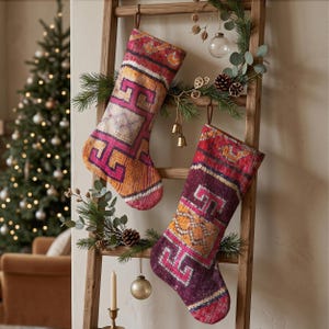 Könnte beinhalten: Zwei Weihnachtsstrümpfe hängen an einer Holzleiter. Der linke Strumpf zeigt ein lebhaftes Muster aus orangefarbenen, rosafarbenen und cremefarbenen geometrischen Formen. Der rechte Strumpf hat ein dunkelviolettes, orangefarbenes und rosafarbenes Design.