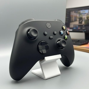 Könnte beinhalten: Ein schwarzer kabelloser Gamecontroller mit farbigen Tasten und Joysticks, der auf einem weißen Ständer steht. Der Controller hat eine matte Oberfläche und ein symmetrisches Design. Der Ständer ist ein einfaches, abgewinkeltes Design.