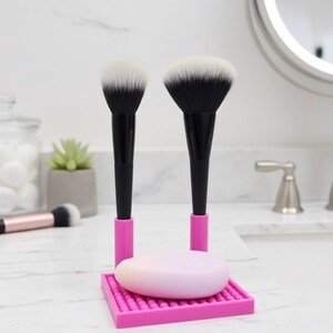 Könnte beinhalten: Zwei Make-up-Pinsel mit schwarzen Griffen und weißen Borsten stehen in einem rosa Halter mit einer rosa Seife. Die Pinsel stehen auf einer weißen Arbeitsplatte in einem Badezimmer. Ein kleinerer Pinsel ist im Hintergrund zu sehen.