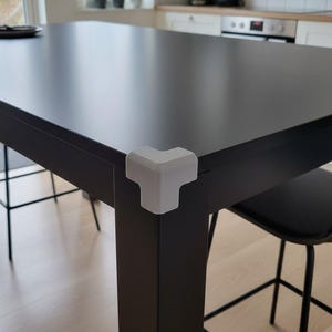 Könnte beinhalten: Ein schwarzer rechteckiger Tisch mit einem weißen Eckenschutz. Der Tisch befindet sich in einer Küche mit Fenster, Pflanzen und einem schwarzen Stuhl. Der Eckenschutz ist hellgrau.