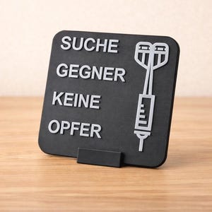 Könnte beinhalten: Schwarzes Schild mit weißem Text und einer Spritzenillustration. Der Text lautet "SUCHE GEGNER KEINE OPFER". Das Schild wird von einem kleinen schwarzen Ständer getragen und steht auf einer Holzoberfläche.