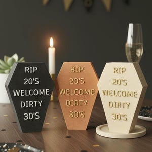 Könnte beinhalten: Drei Sarg-förmige Dekorationen mit dem Text "RIP 20's WELCOME DIRTY 30's". Die Dekorationen sind schwarz, bronze und cremefarben. Eine brennende Kerze und ein Sektglas sind im Hintergrund. Auf einem Holztisch.