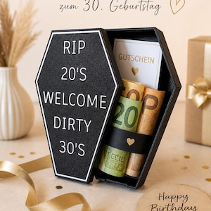 Könnte beinhalten: Eine schwarze Sarg-Geschenkbox mit weißem Text "RIP 20's WELCOME DIRTY 30's". Im Inneren befinden sich gerollte Banknoten und eine Geschenkkarte. Die Box steht auf einem Tisch mit einer "Happy Birthday"-Karte und goldenem Band.