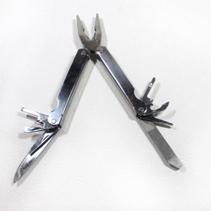 May include: A multi-tool with a silver finish, featuring pliers, a knife, a file, and other tools. The tool is open, displaying its various components against a white background. The pliers are at the top, and the file is at the bottom.