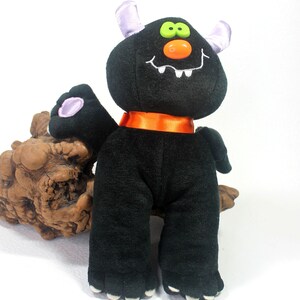 May include: A black plush monster toy featuring two light purple horns, bright green eyes, an orange plastic nose, and a wide white embroidered mouth with teeth. The creature wears a smooth orange ribbon collar and has purple paw pads on its feet.