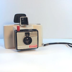 May include: A vintage Polaroid Swinger Model 20 Land Camera, beige and black, with a red button and a black wrist strap. The camera has a square design with a silver-colored lens and flash attachment.
