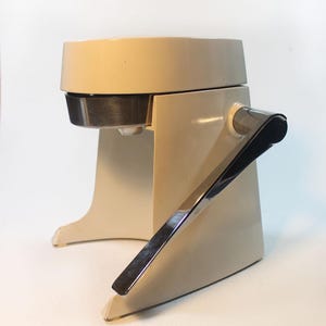 May include: A vintage cream-colored citrus juicer with a stainless steel strainer and a chrome handle. The juicer has a classic design with a wide base and a curved handle for easy use. The juicer is made of durable materials and is designed for efficient juice extraction.