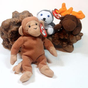 May include: A brown plush monkey, a white and gray plush husky, and a brown plush moose with orange antlers are displayed. The toys have tags attached. The toys are on a brown, textured base.